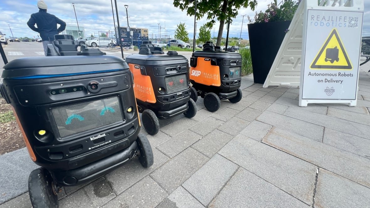 skip-robots-in-markham-by-real-life-robotics-1 Three orange robots in a row with a sign beside that reads: Autonomous Robots on Delivery.