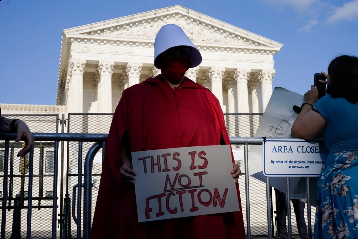 supreme-court-abortion A woman wearing a red rob and large white bonnet stands outside a large white courthouse holding a sign that reads "This is not fiction."