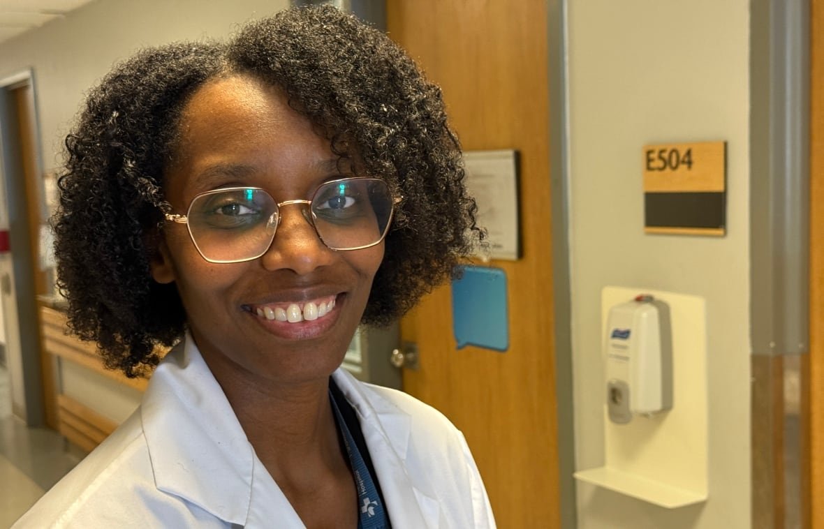 sybile-delice-charlemagne-may-26-2025 A woman in a white lab coat with an Ottawa Hospital lanyard stands next to a hand sanitizer disepenser.