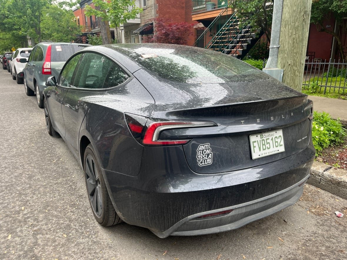 tesla-montreal A grey EV car with a bumper sticker is parked on a residential street.
