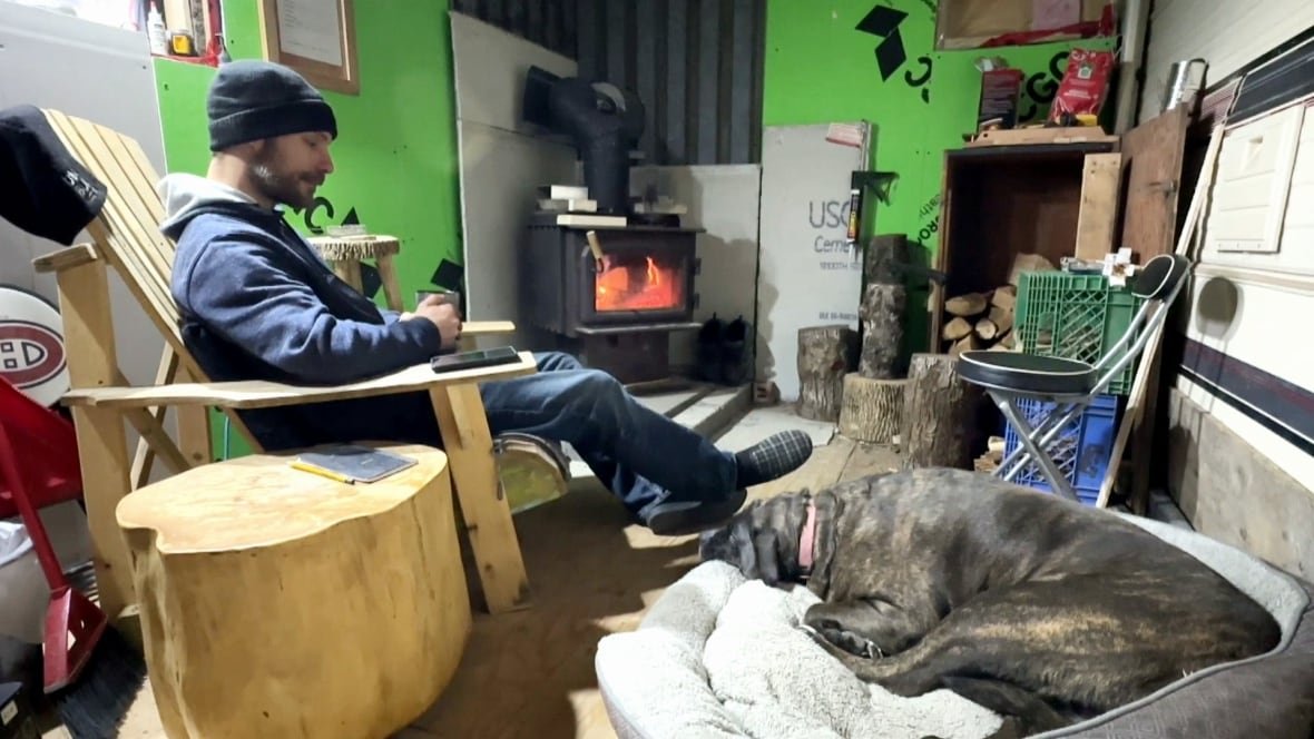 tetreault-interior-rv A white man in a blue sweater and black knit hat sits in a wooden yellow chair with a mug in his hand, a large dark brown dog laying on a cushion at his feet. There is a wood stove in the background