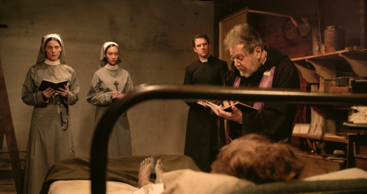 the-ritual-2 A priest reading from a book stands in front of a woman in a bed and beside two nuns and another priest.. Only the woman's feet are visible, which are bare and dirty.