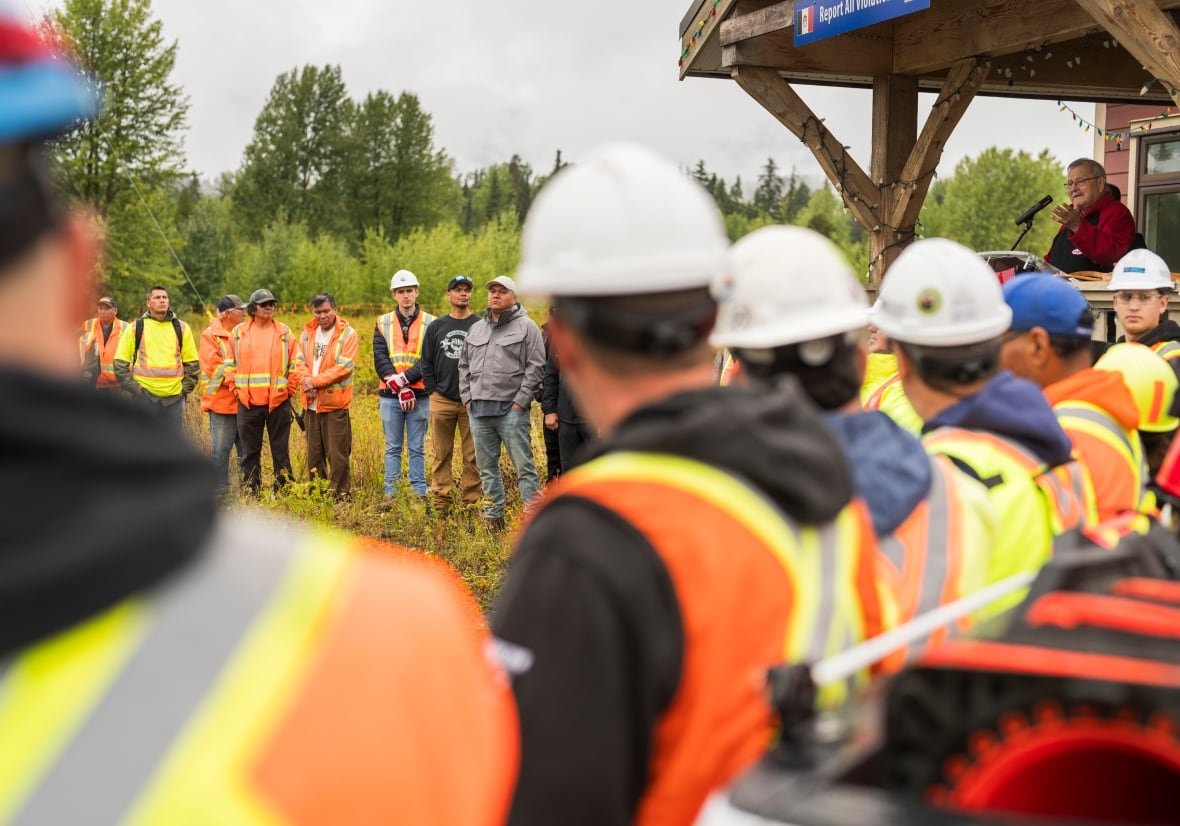 western-lng-pipeline-ceremony Men in hard hats and construction vests stand in a row listening to an Indigenous Elder.
