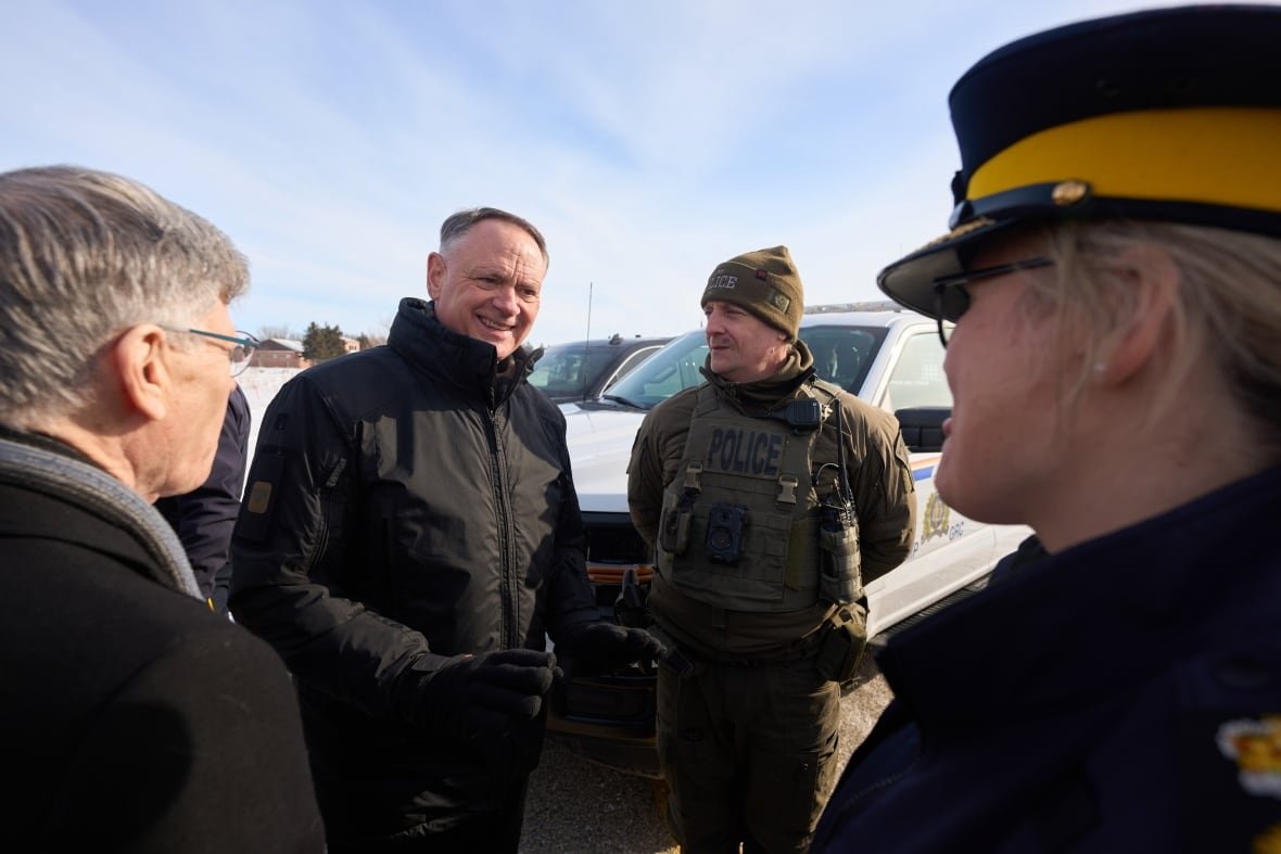 Two men in winter jackets stand in front of a car, casually chatting with two uniformed police officers.