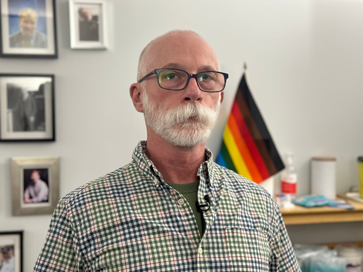 chris-aucoin a man wearing glasses and a button up shirt looks into the canera. A pride flag and portrait are in the background.