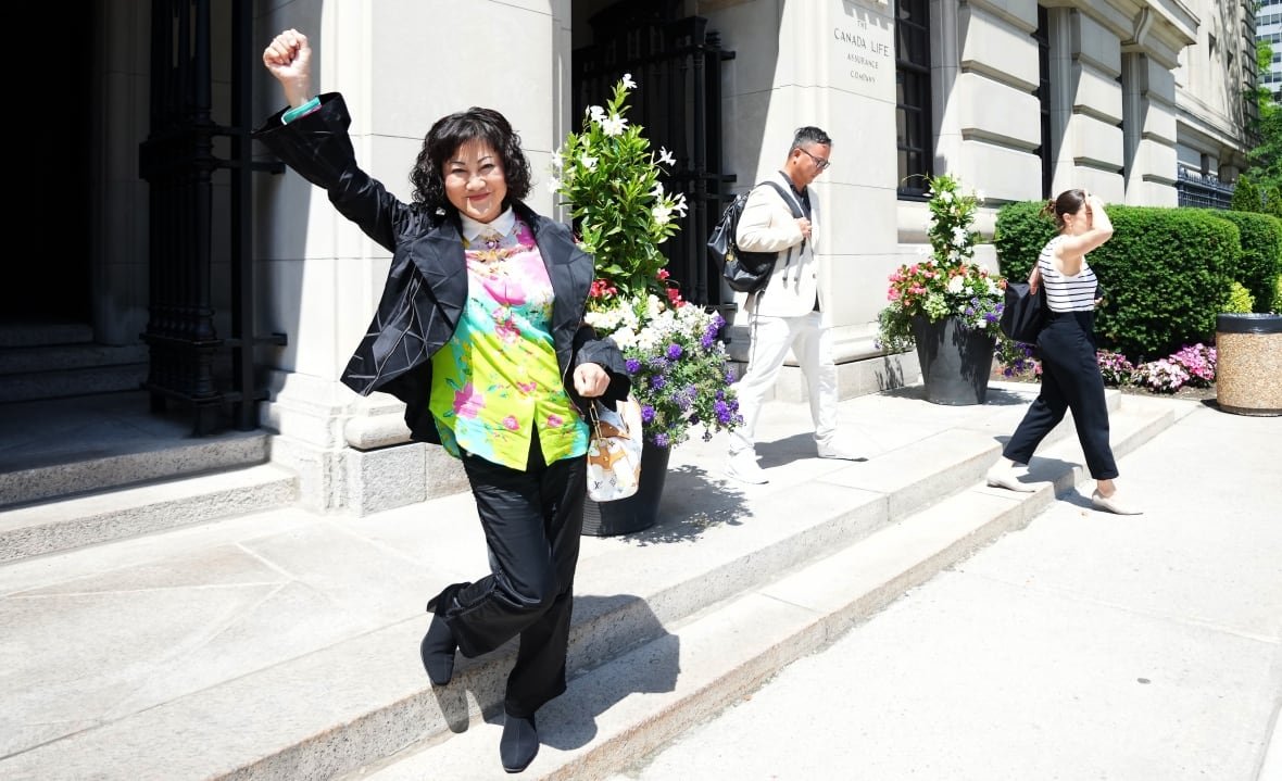 hudsons-bay-ccaa-20250623 A woman with a black bob, black jacket, floral shirt and black pants pumps a fist in the air, standing on the steps of a courthouse.