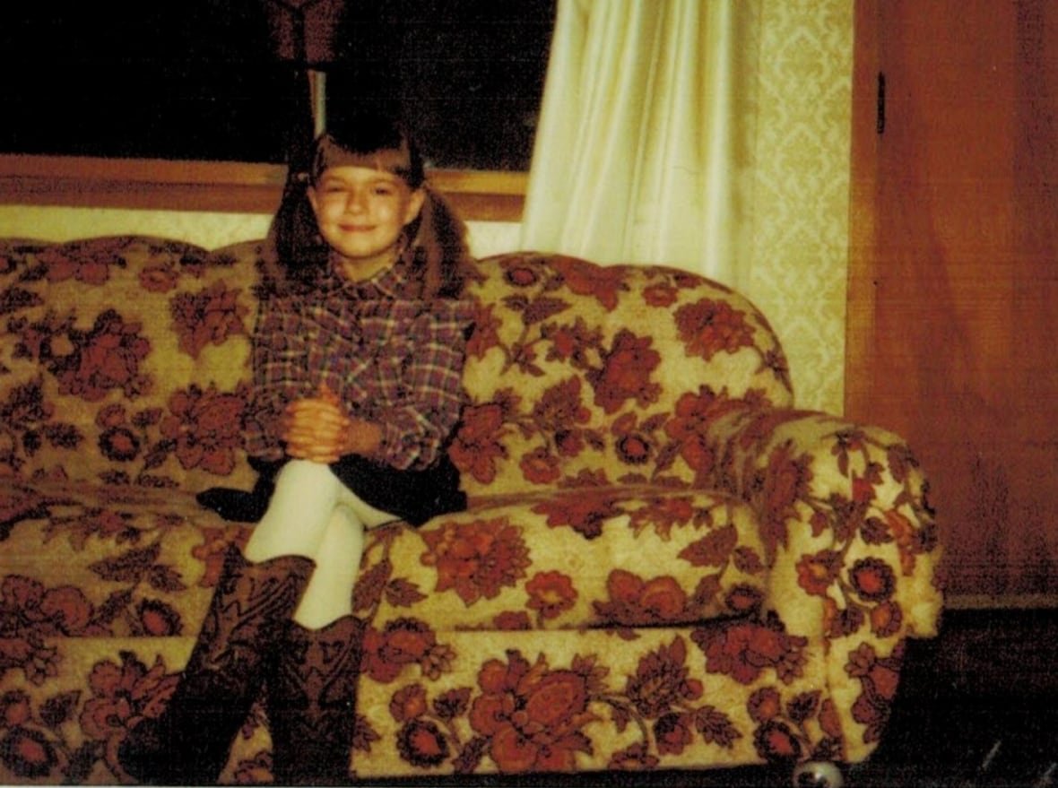 jennifer-lovegrove-child A smiling pigtailed pre-teen girl sits on a flowered couch wearing a western themed plaid shirt and oversized cowboy boots.