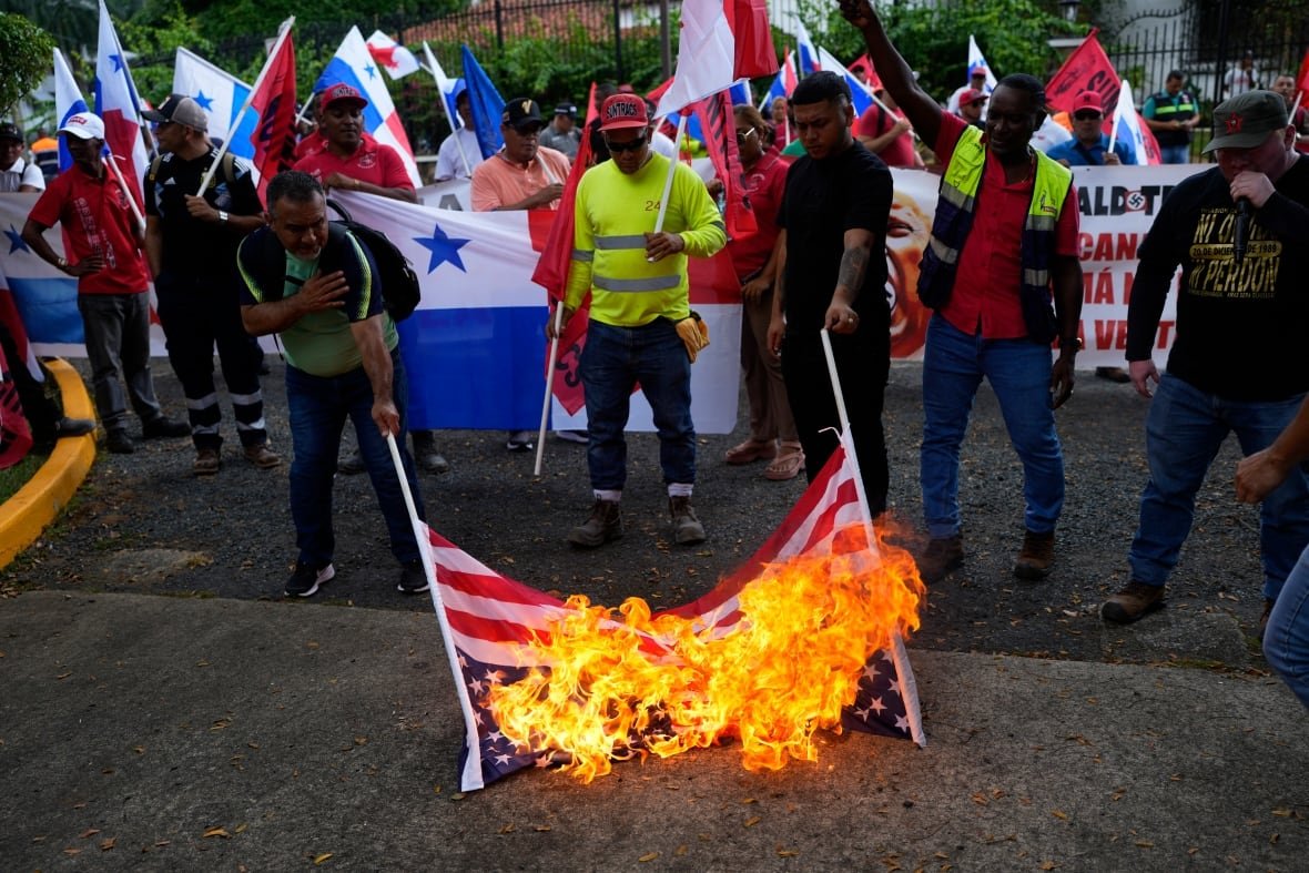 panama-protest A group of protesters holding flags rally outside a residence. In the foreground, a flag burns on the ground in front of a man wearing a yellow safety vest.