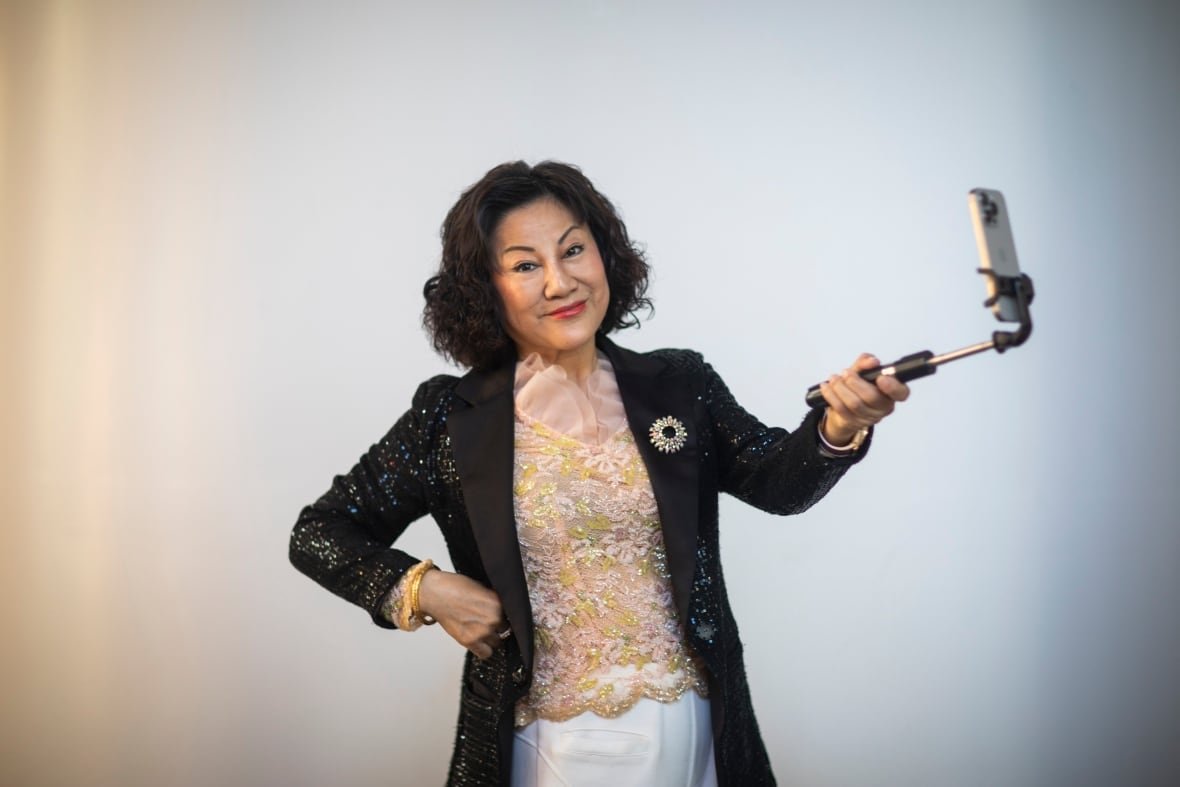 ruby-liu-1 An East Asian woman with a dark bob haircut smiles while holding a selfie stick. She wears a sparkly pink and green top with a sparkly black blazer over top.