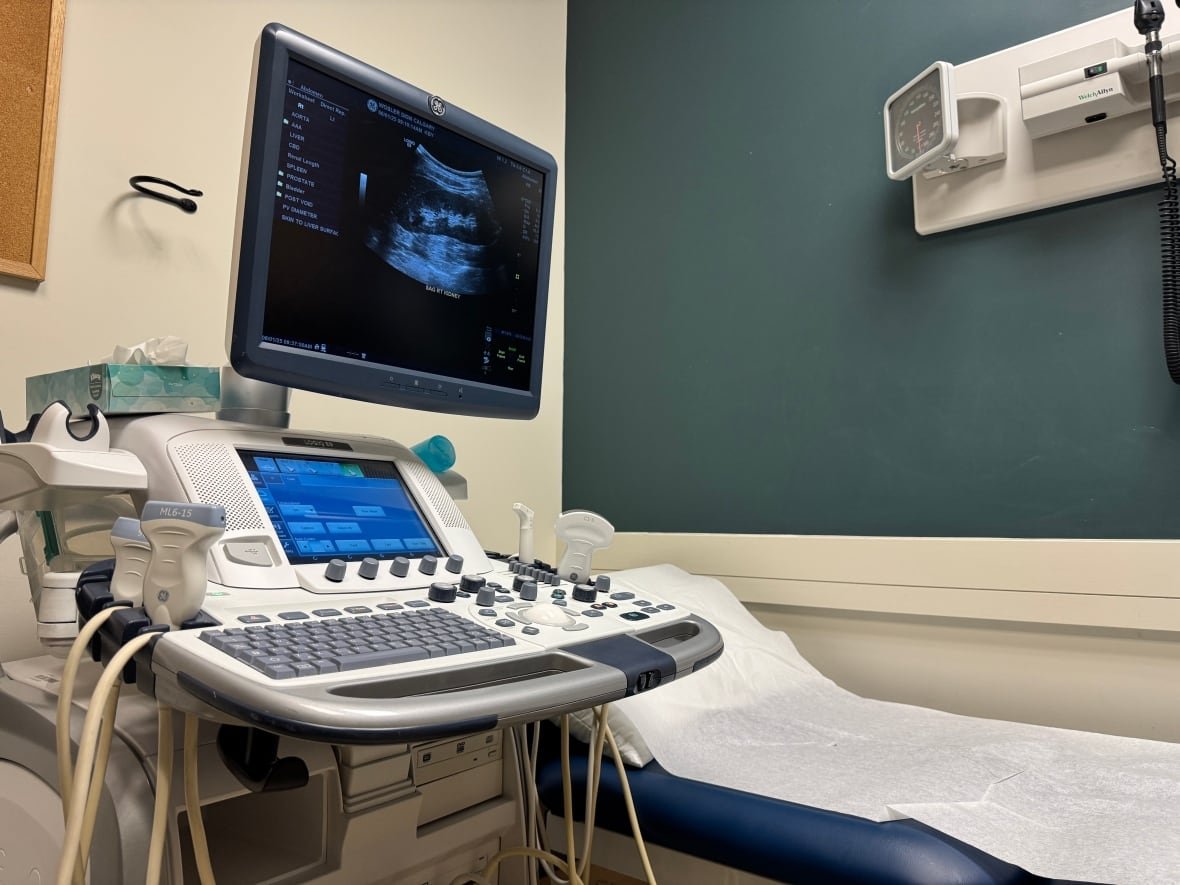 wosler-ultrasound-clinic An ultrasound machine next to an empty patient bed. The monitor displays an ultrasound image of an abdomen.