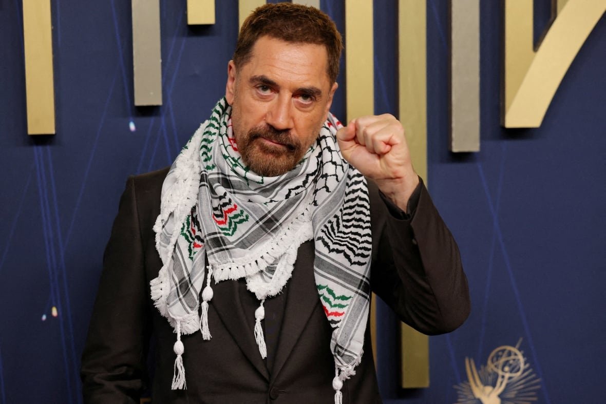 a middle aged man with dark hair and wearing a black suit and a black and white scarf holds his fist up in the air on a red carpet