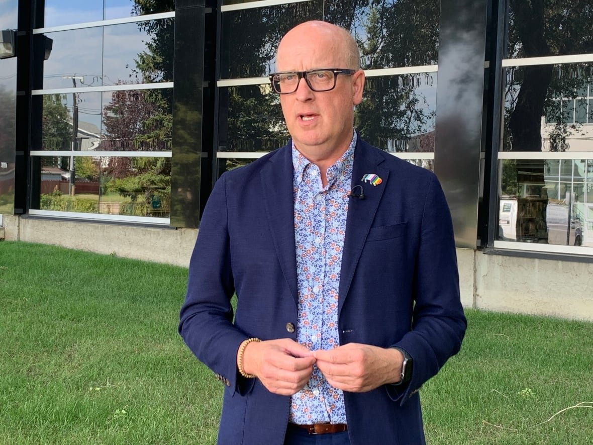 A man in a navy blazer and flowered dress shirt stands in front of a glass building on a green lawn.