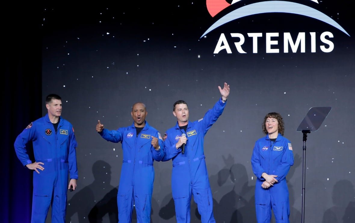 nasa-moon-astronauts Four astronauts in blue outfits stand together, some mid-whoop, others smiling. An Artemis logo is visible on the wall behind them.