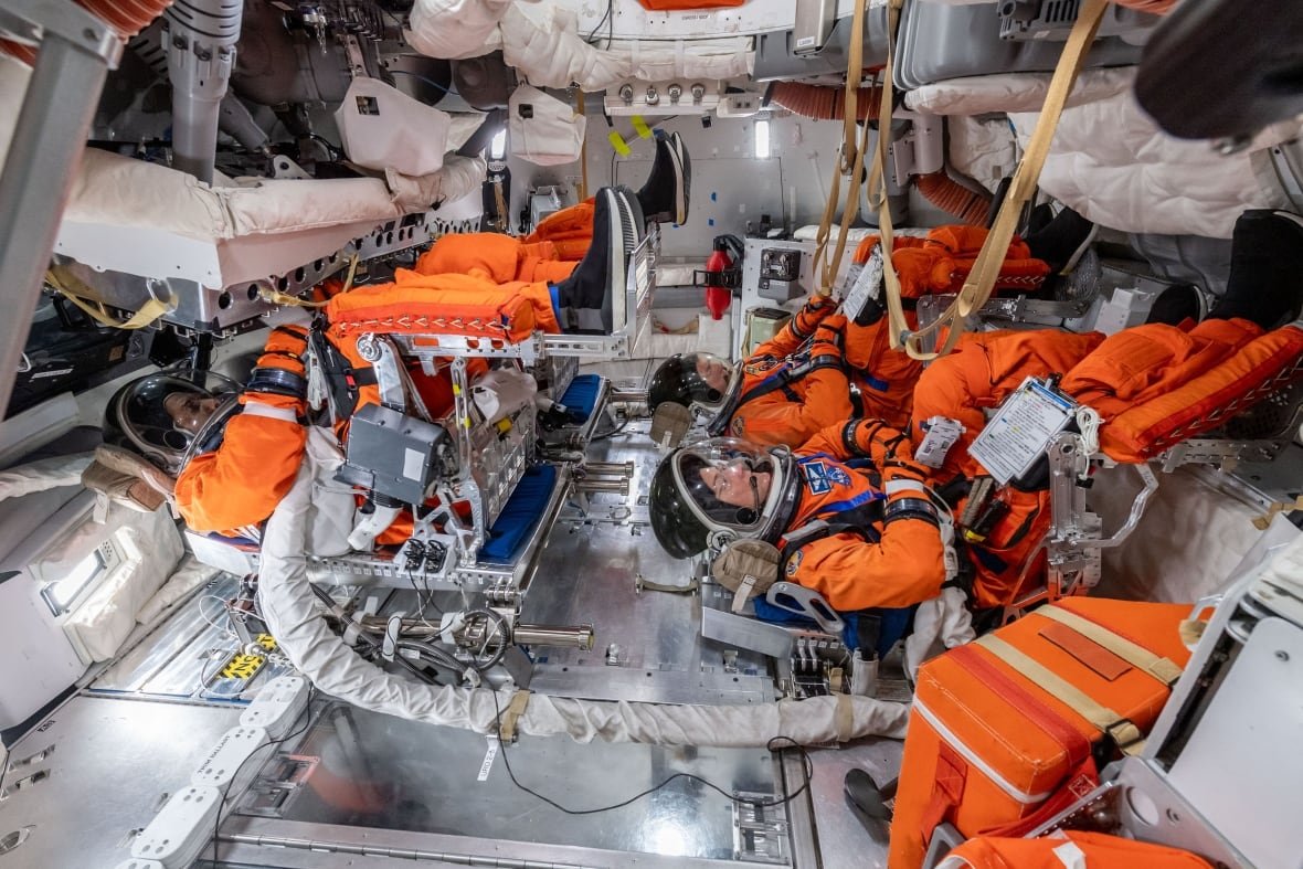 orion-simulator-artemis-ii Four people in orange spacesuits are seen lying in a spacecraft.