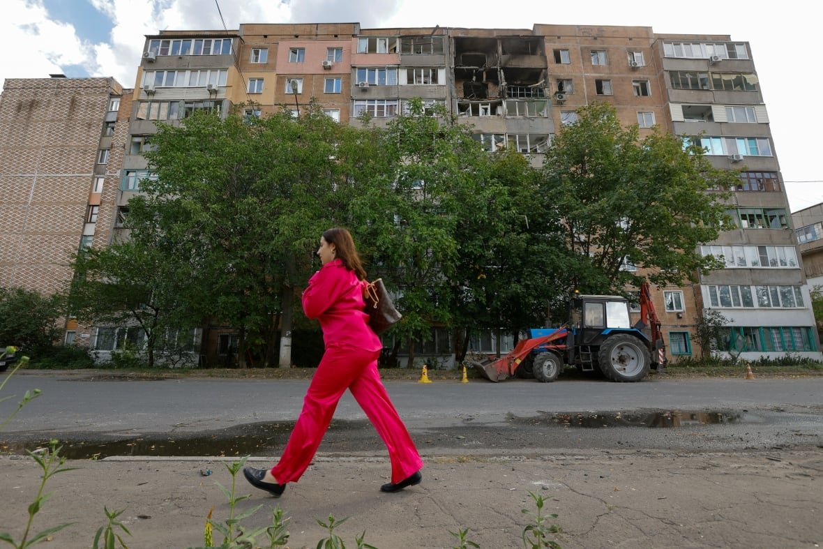 ukraine-crisis-makiivka-attack A woman wearing all red walks on a sidewalk in the foreground with a six-storey residential building across the street with heavy damage to its exterior, including damaged balconies and windows.
