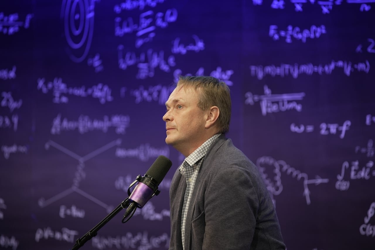 default-948 A man wearing a dark gray suit sits in front of a podcast microphone.