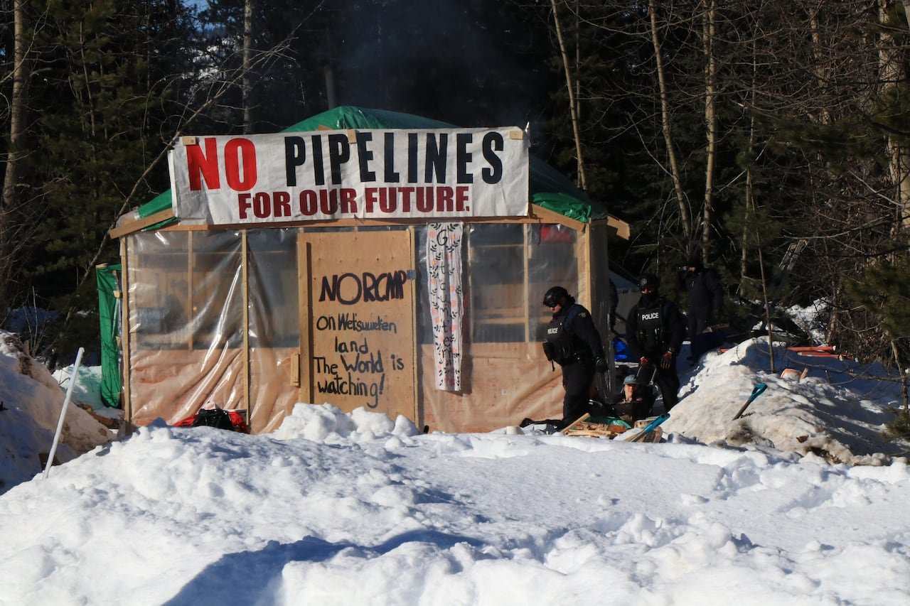 default-290 A makeshift hut constructed of wood and tarps beside a snowy forest, with three police officers passing by. A sign on top of the hut reads: No pipelines for our future.