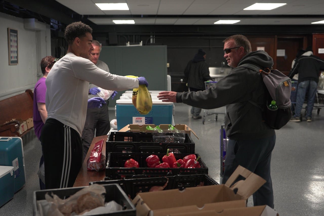 A young man hands a bag to an older man wearing sunglasses and a coat in what appears to be a transaction. The two people are separated by a table with fruits and vegetables on it.