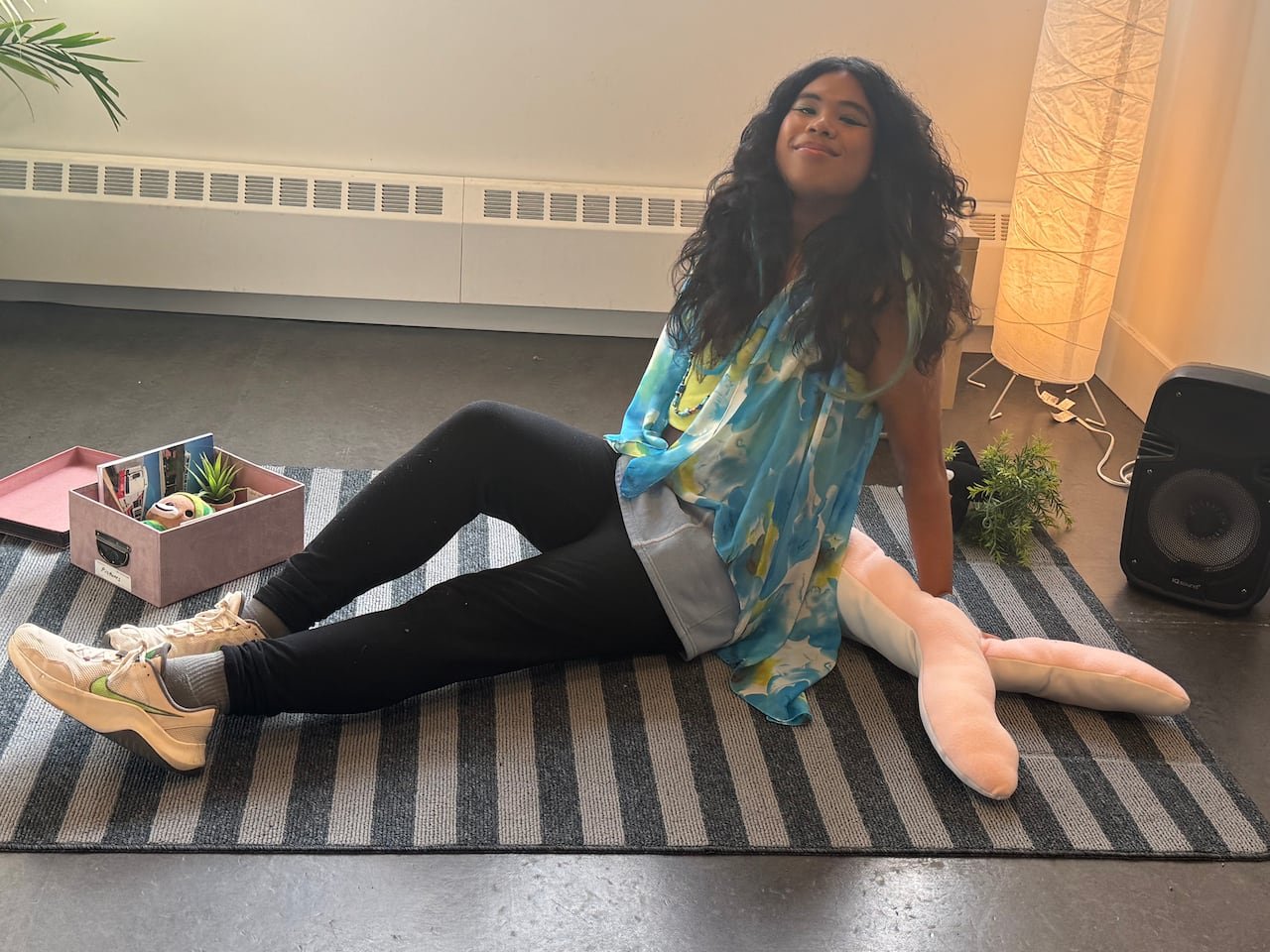 A smiling queer youth with curly long dark hair sitting on a carpet