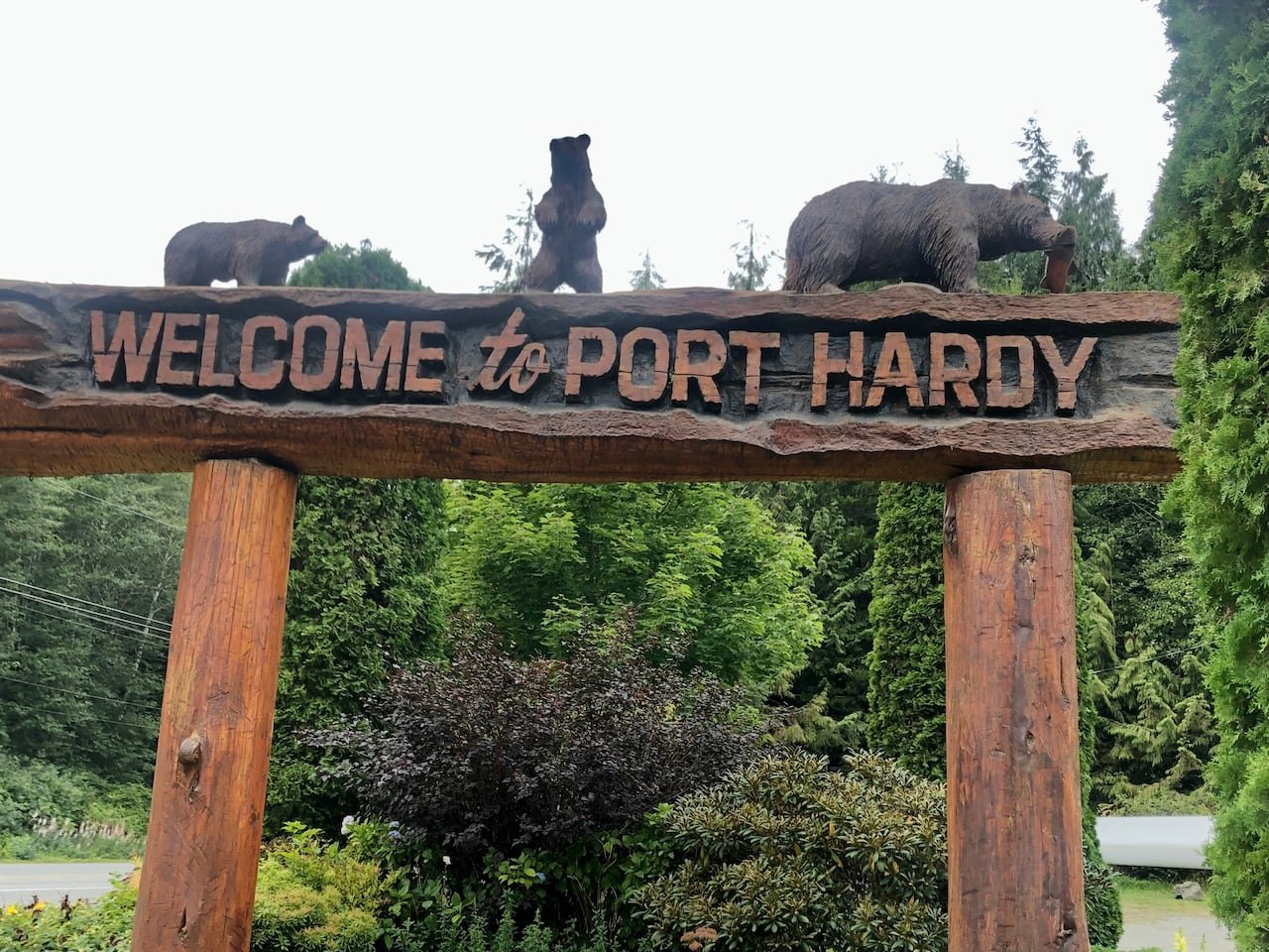 A sign carved out of logs reads "Welcome to Port Hardy." On top there are three carved bears.