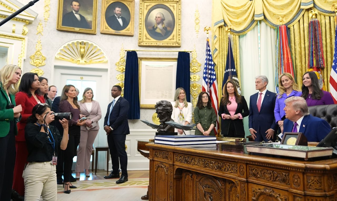 default-167 A wide-angle photo in the Oval Office shows Donald Trump sitting at his desk, looking across toward a group of reporters, with gold-framed paintings of presidents on the wall behind.