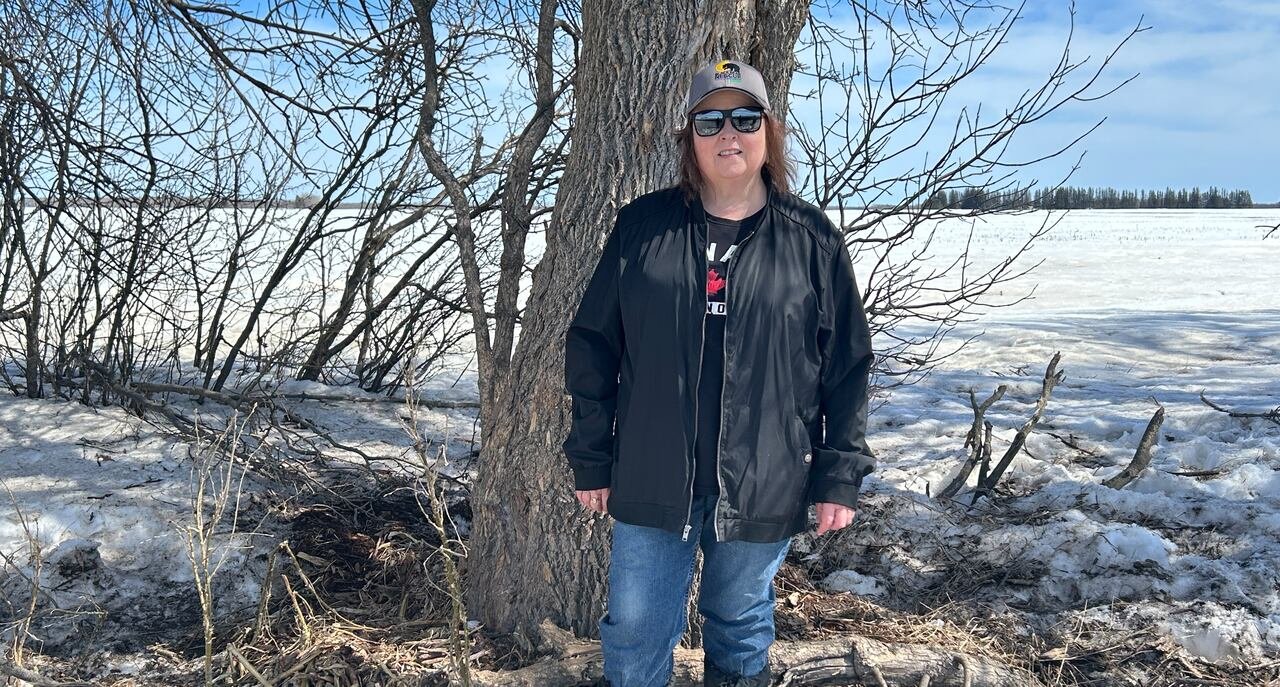 default-93 Woman wearing black spring jacket, blue jeans, a baseball cap and sunglasses while standing in front of a tree with snow on the ground